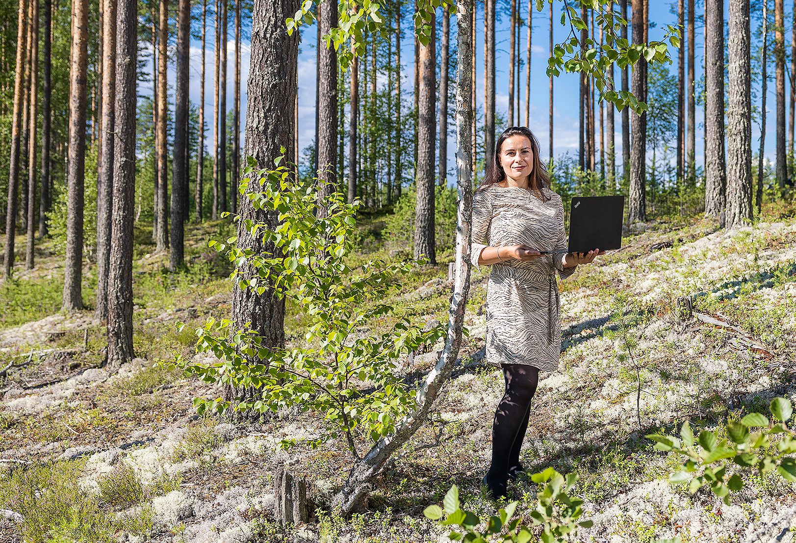 Askolan kunnanjohtaja Annu Räsänen seisoo metsän keskellä kannettava tietokone kädessä. Blogitekstin kuvituskuva