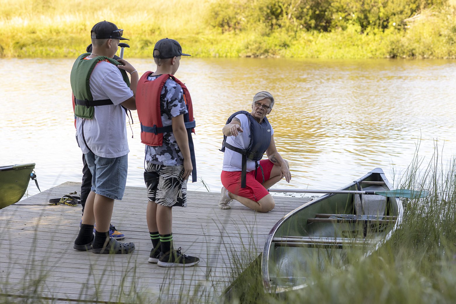 nuoriso-ohjaaja opastaa joen rannalla kolmea nuorta astumaan kanoottiin. kaikilla kesäinen vaatetus ja pelastusliivit päällä